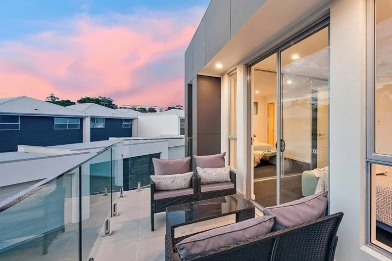 Balcony with outdoor seating, including two armchairs and a sofa, overlooking neighboring modern houses at sunset.