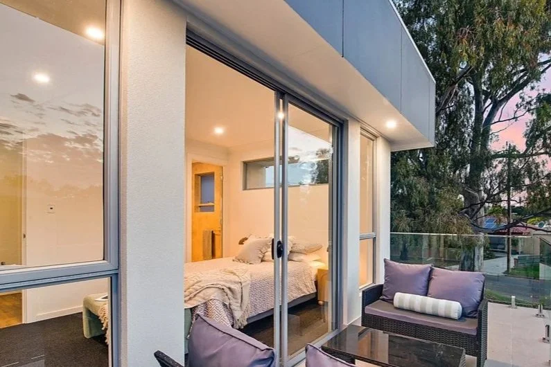 View of a modern bedroom through glass sliding doors, with outdoor patio furniture, trees, and a sunset sky in the background.