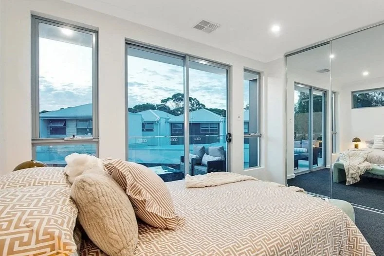 A modern bedroom with large windows showing a view of neighboring houses and a balcony, with a bed dressed in beige and white geometric patterned bedding and multiple pillows.