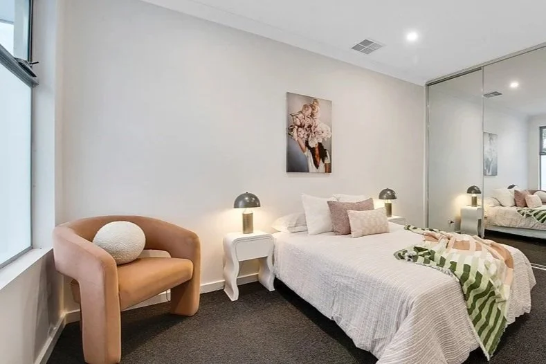 A bedroom featuring a bed with multiple pillows, a side table with a lamp, a peach-colored armchair with a round pillow, a piece of floral artwork on the wall, and a mirrored closet door reflecting part of the room.
