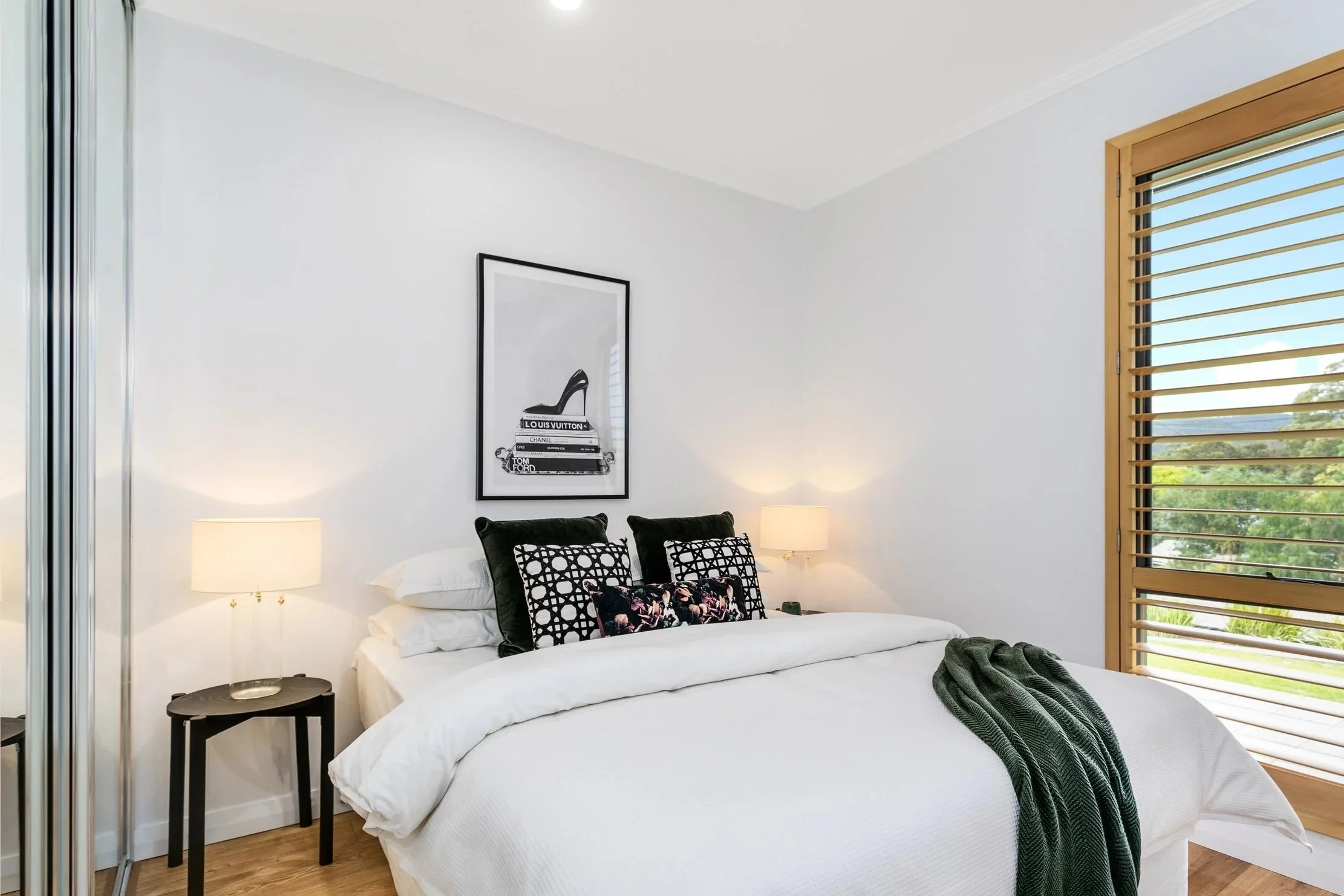 A modern bedroom with a white bed, black and patterned pillows, two bedside lamps, a warm wood window shutter, and framed black and white artwork of books and a high heel shoe.