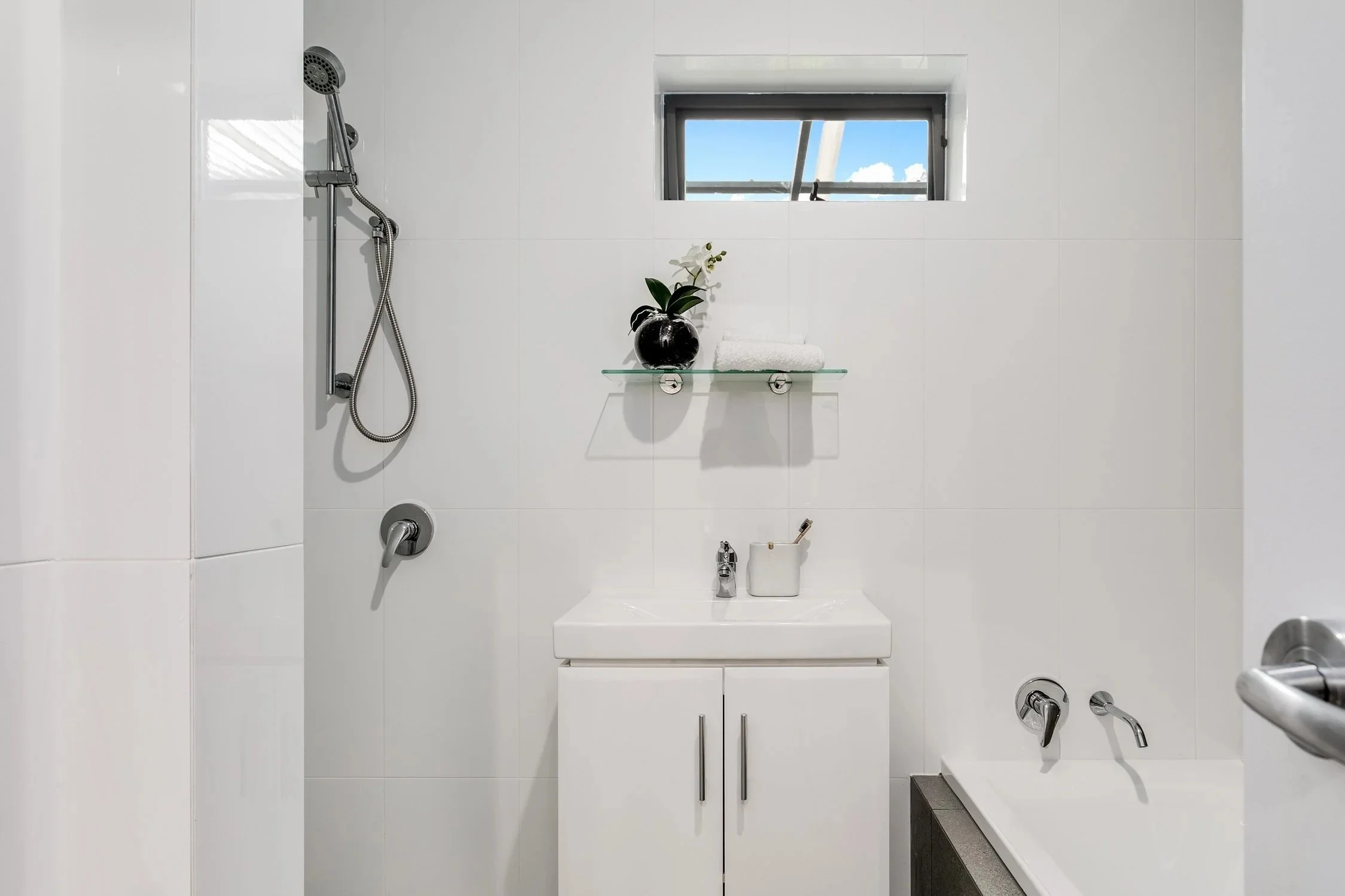 Minimalist white bathroom with a small window, sink, black vase with white flower, towel on shelf, shower with handheld head, and bathtub.