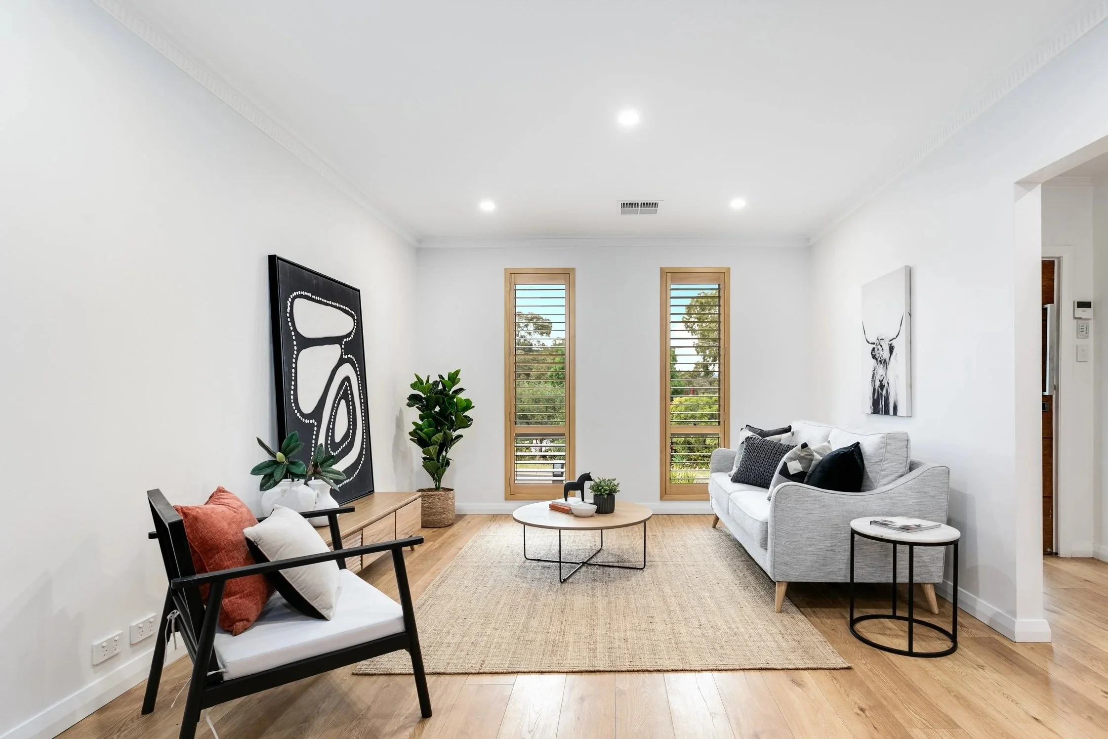 Modern living room with white walls, light wood floors, two vertical windows with wooden frames, a light gray sofa with black and white pillows, a black armchair with white and rust pillows, a rectangular beige rug, a round coffee table with small de