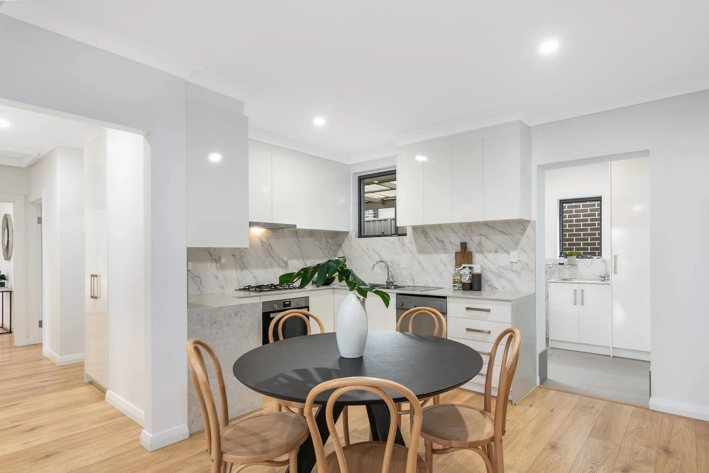 Modern kitchen with white cabinets, marble backsplash, and a round black dining table with wooden chairs, featuring a white vase with green leaves at the center.
