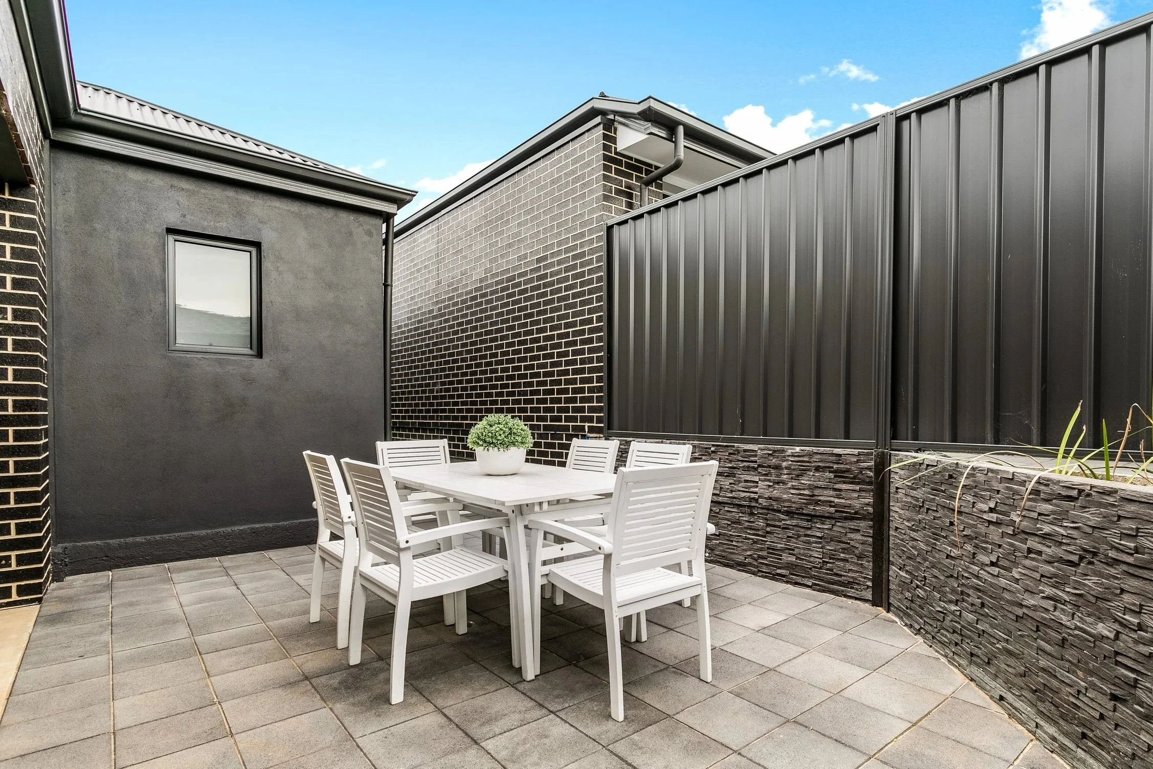Outdoor patio with a white table and six matching chairs, potted plant in the center, surrounded by black brick and metal fence walls, under a partly cloudy sky.