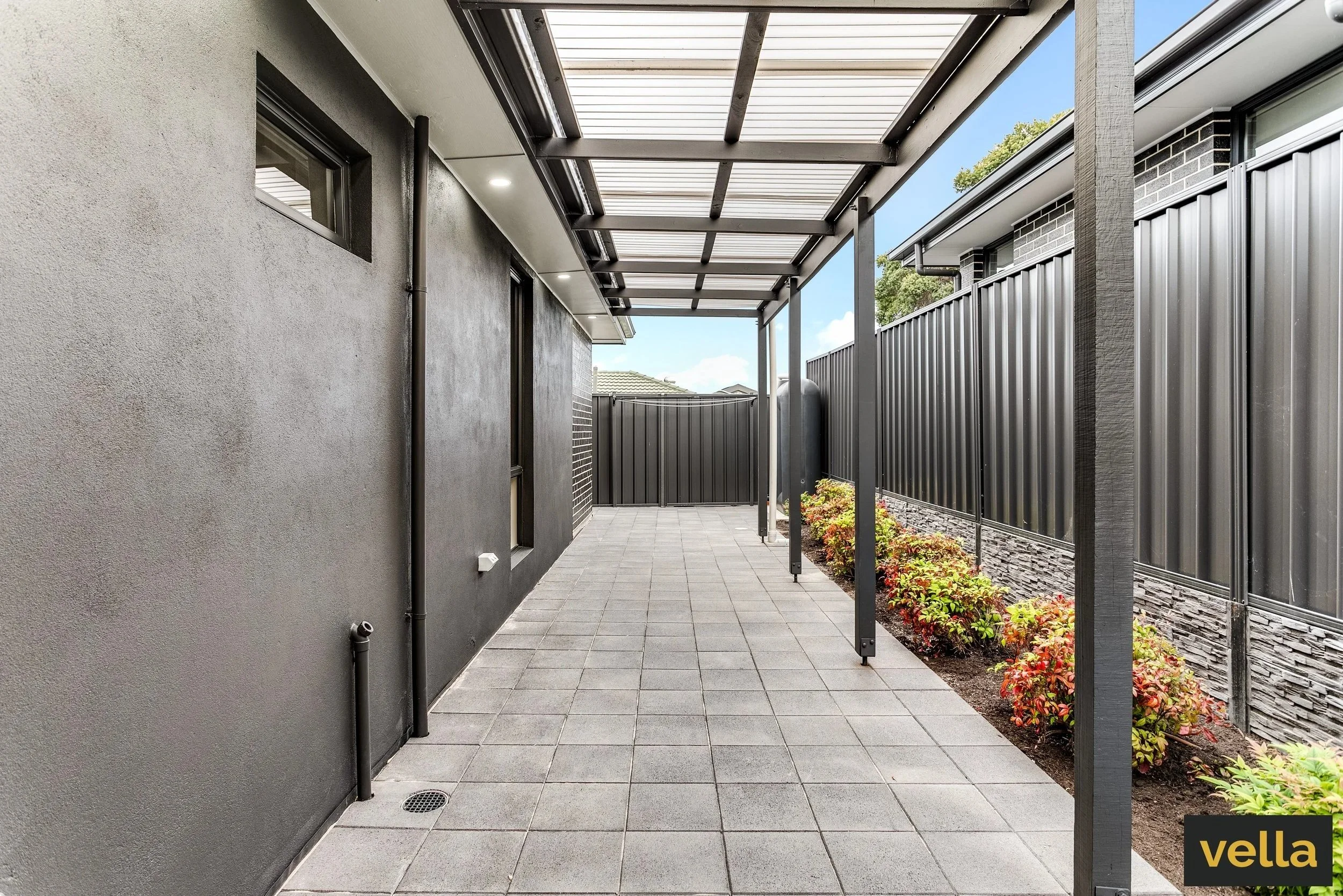 A modern backyard patio with gray pavers, a protective metal roof, black side walls, and some small plants along the fence.