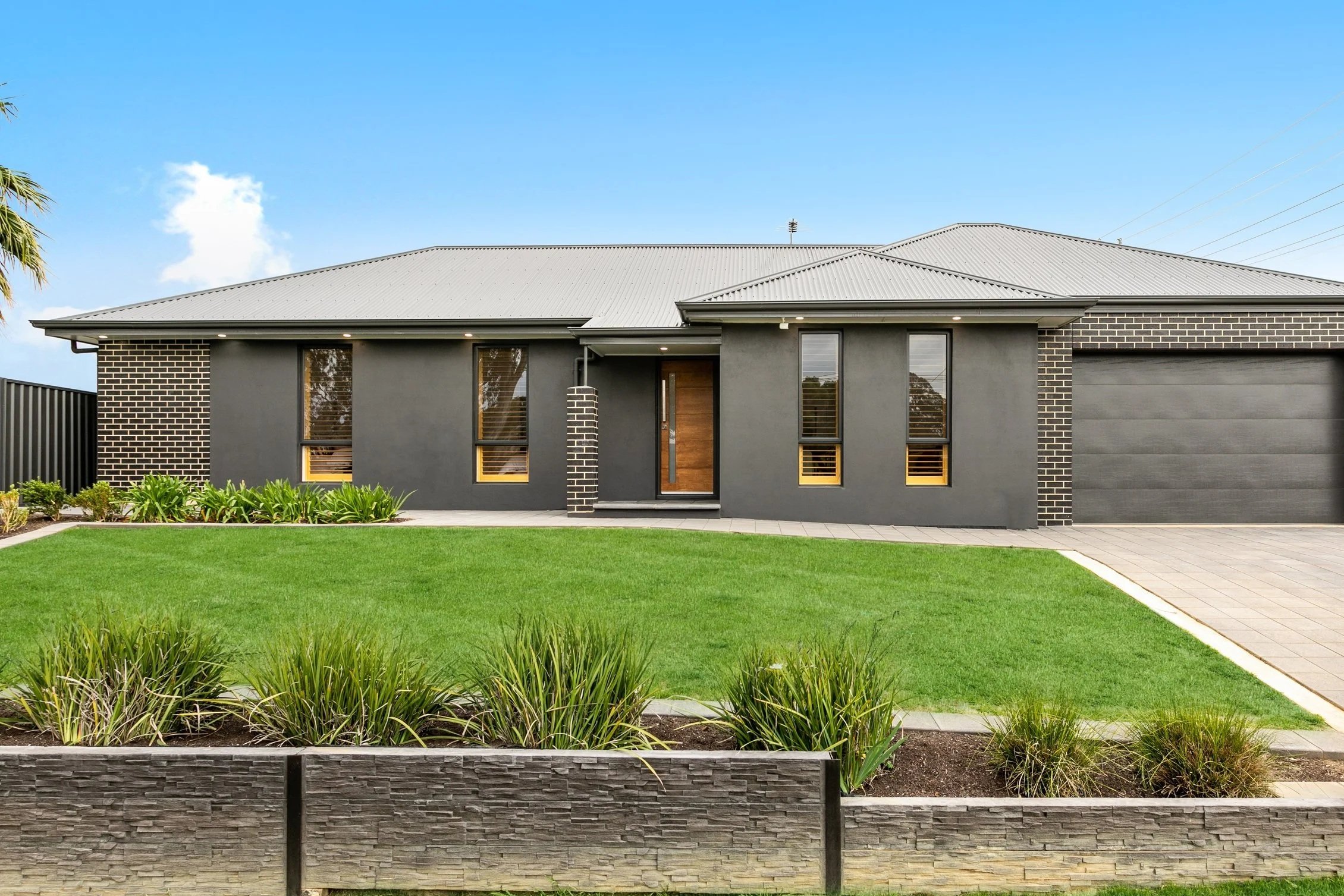 Modern single-story house with a gray facade, brick accents, and a metal roof, surrounded by a green lawn and landscaped garden.