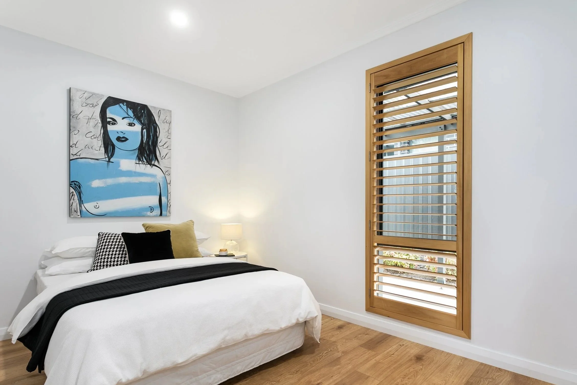 A bedroom with a white bed, black and gray pillows, a small lamp on nightstand, a window with wooden slats, and a artwork of a woman with dark hair and blue skin hanging on the wall.