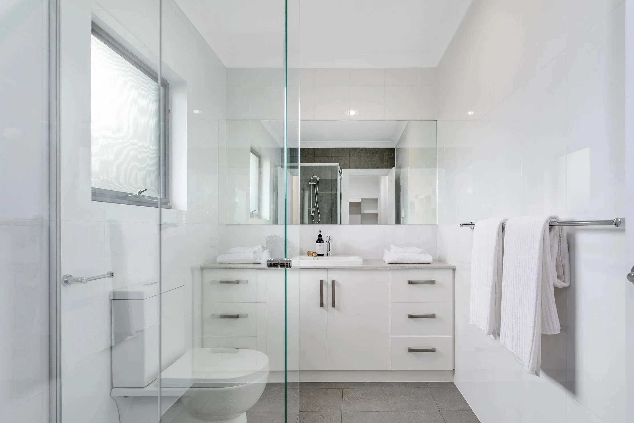 Clean, modern bathroom with white cabinetry, large mirror, towels, and a window