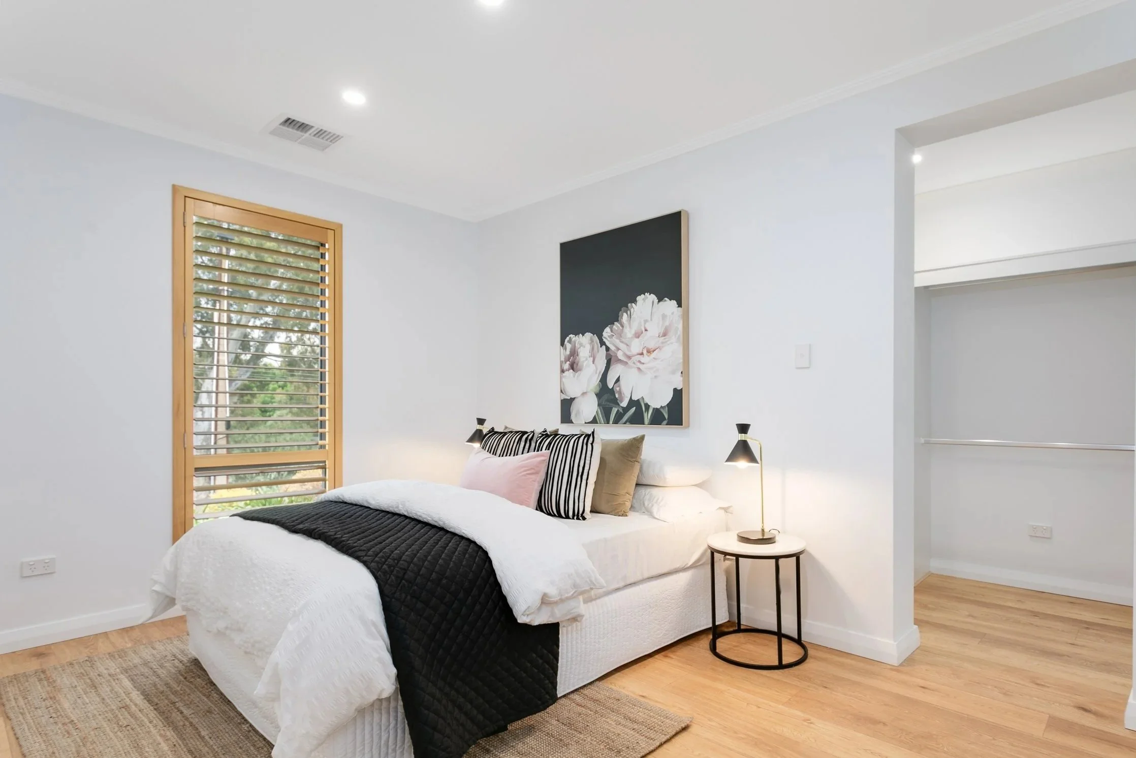 A modern bedroom with white walls, a wooden window with blinds, a bed with white bedding and black, pink, and beige pillows, a nightstand with a black lamp, a large floral artwork above the bed, and a walk-in closet with a railing.