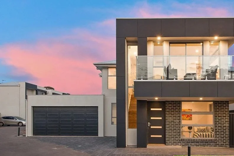 Modern two-story house with large glass windows and balcony, black and brick exterior, sunset sky in background.