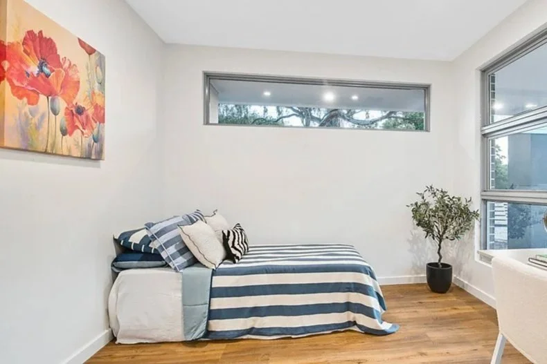 A minimalist bedroom with a single bed covered in blue and white striped bedding, a colorful floral painting on the wall, a potted plant near the window, and large windows providing natural light.