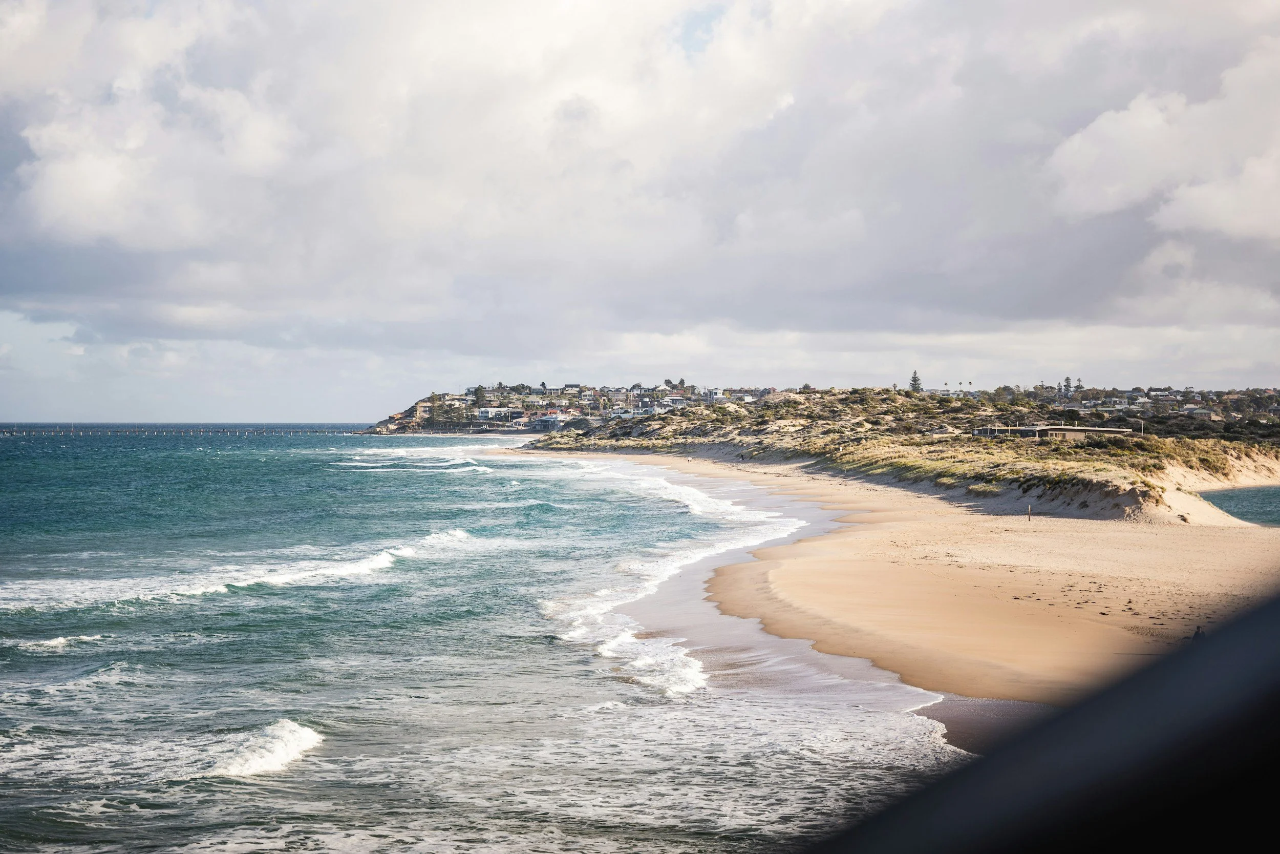 A coastal scene with a sandy beach, ocean waves, and a residential area on a hill in the background under cloudy skies.