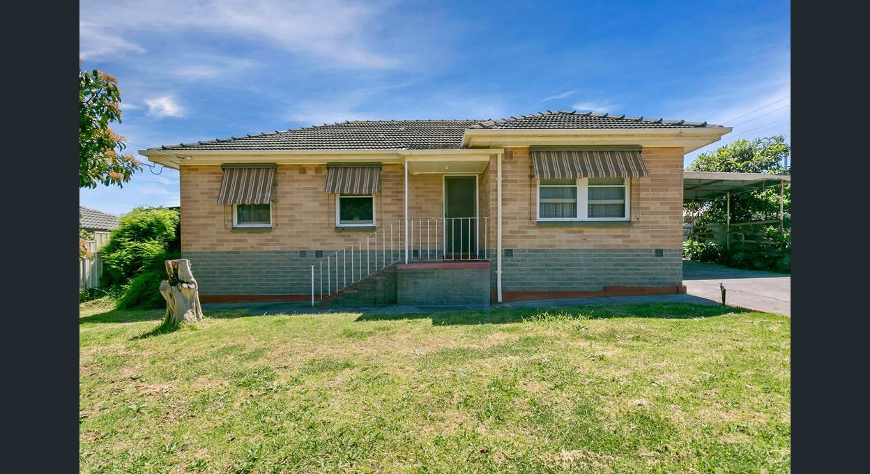 A single-story brick house with brown brick siding and a gray tiled roof, featuring two windows with striped awnings, a front door, and a small front porch with stairs, set against a grassy yard and blue sky.