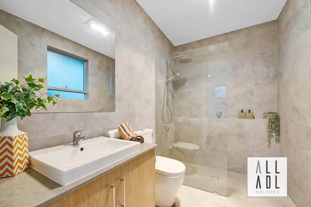 Modern bathroom with beige tiled walls, a white rectangular sink with a wooden cabinet below, a large mirror, a small frosted window, a toilet, and a walk-in shower with a glass partition and a rain shower head. There is a plant and toiletries on the shelf.