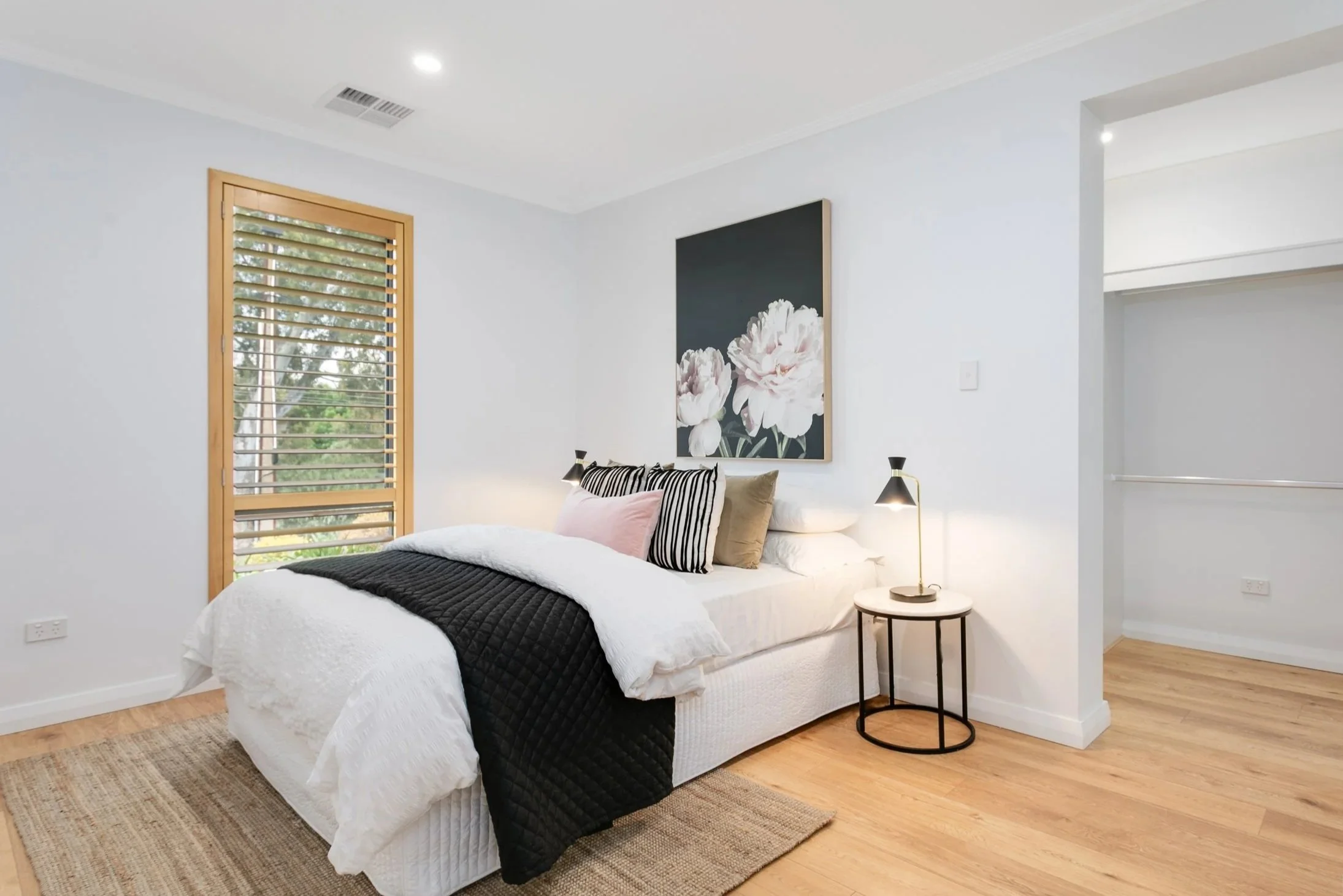 Bright bedroom with white walls, wooden floor, window with slatted blinds, bed with white bedding, black quilt, pink, black-and-white, and tan pillows, artwork of white flowers above the bed, small round side table with a black lamp.