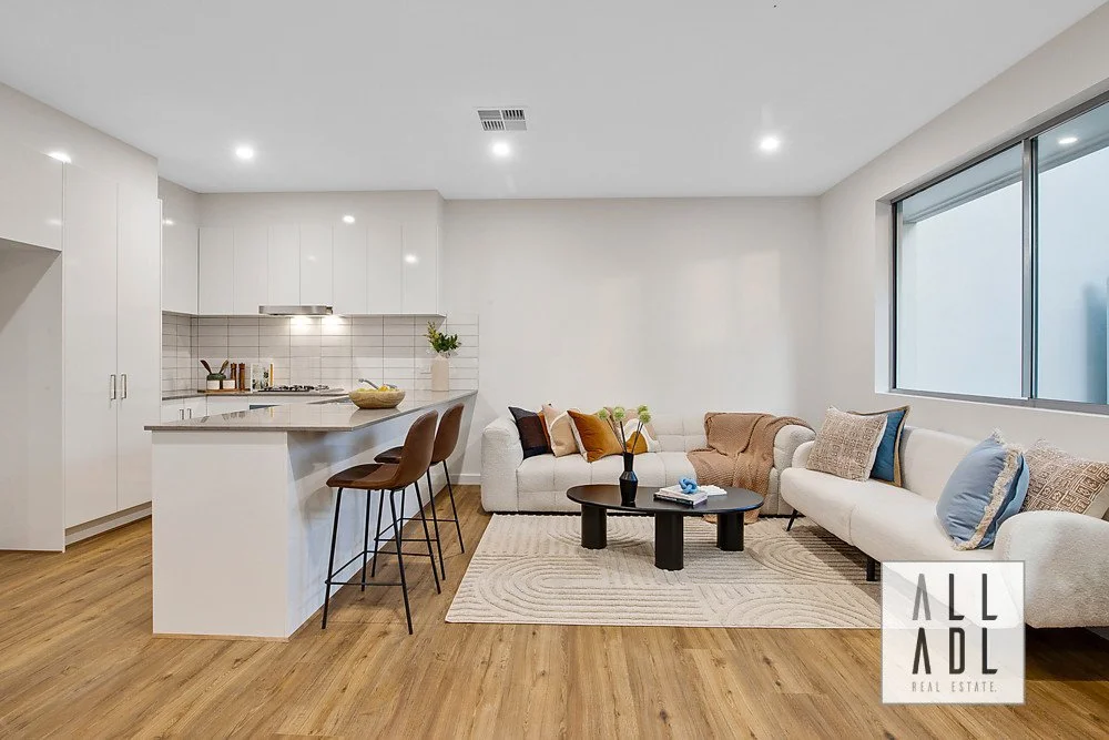 Modern open-plan living room and kitchen with white cabinetry, brown bar stools, beige sofa, coffee table, large window, and hardwood flooring.