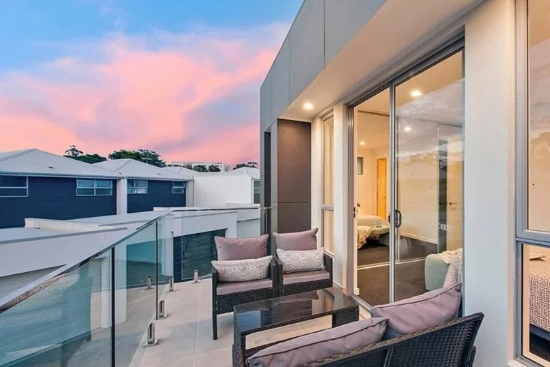 An outdoor balcony with wicker furniture and cushions next to a glass sliding door leading into a bedroom, overlooking neighboring rooftops during sunset.