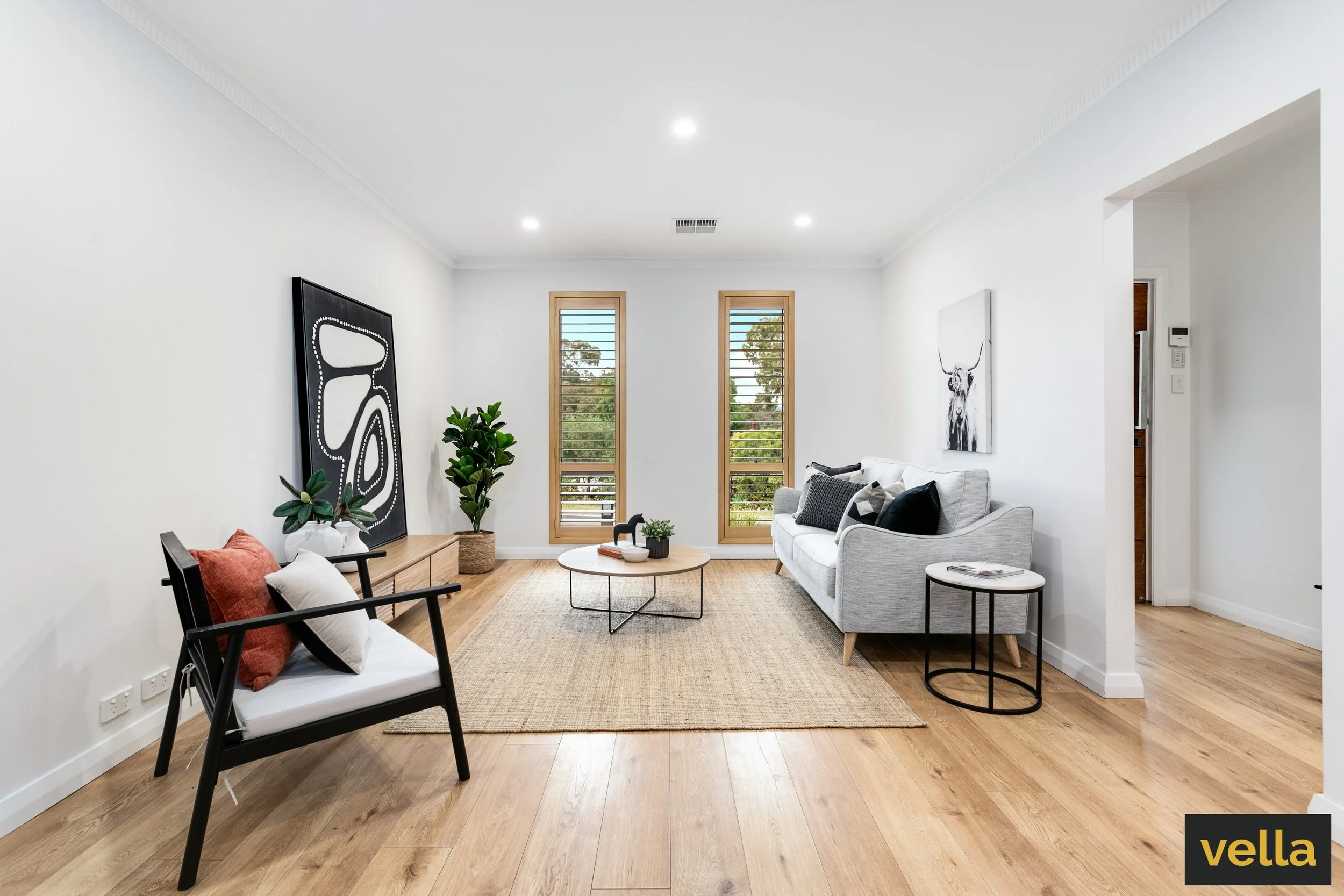 Modern living room with white walls, wooden flooring, a gray sofa with black and white pillows, a black and tan armchair with pillows, a round coffee table, and artwork on the walls, illuminated by natural light from two tall windows.