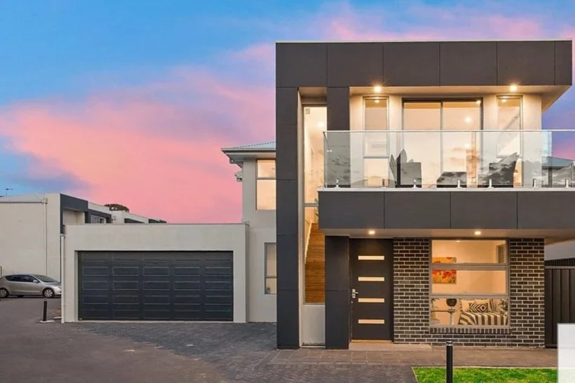 Modern two-story house with a garage, glass balcony, and large windows at sunset.