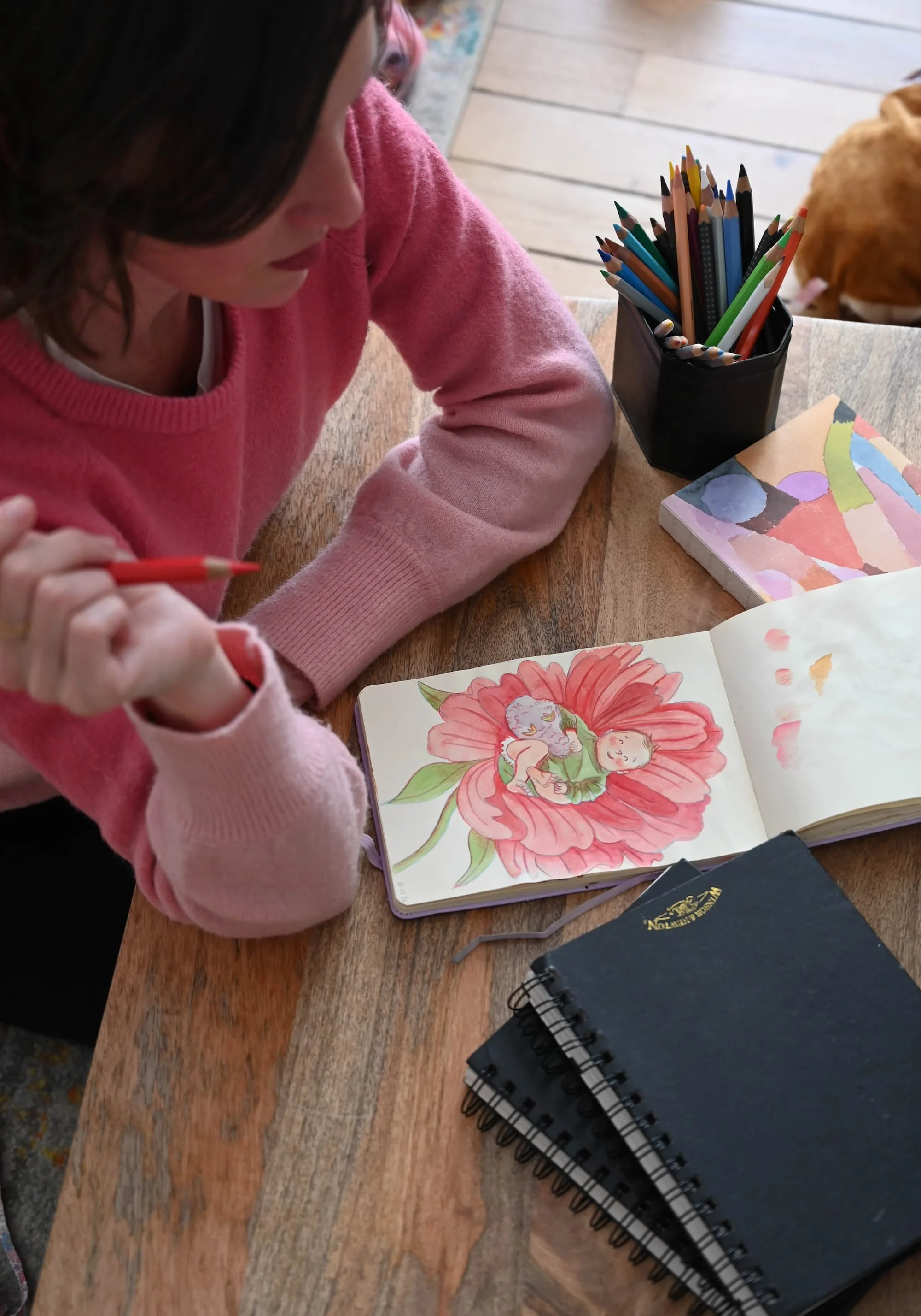 Une jeune fille en pull rose est assise à une table en train de dessiner ou peindre dans un carnet de croquis avec une illustration d'une fille dans une fleur. Sur la table, il y a un jeu de crayons de couleur dans un vase, plusieurs carnets à spirale, et une boîte de peinture ou un cahier avec un fond coloré.