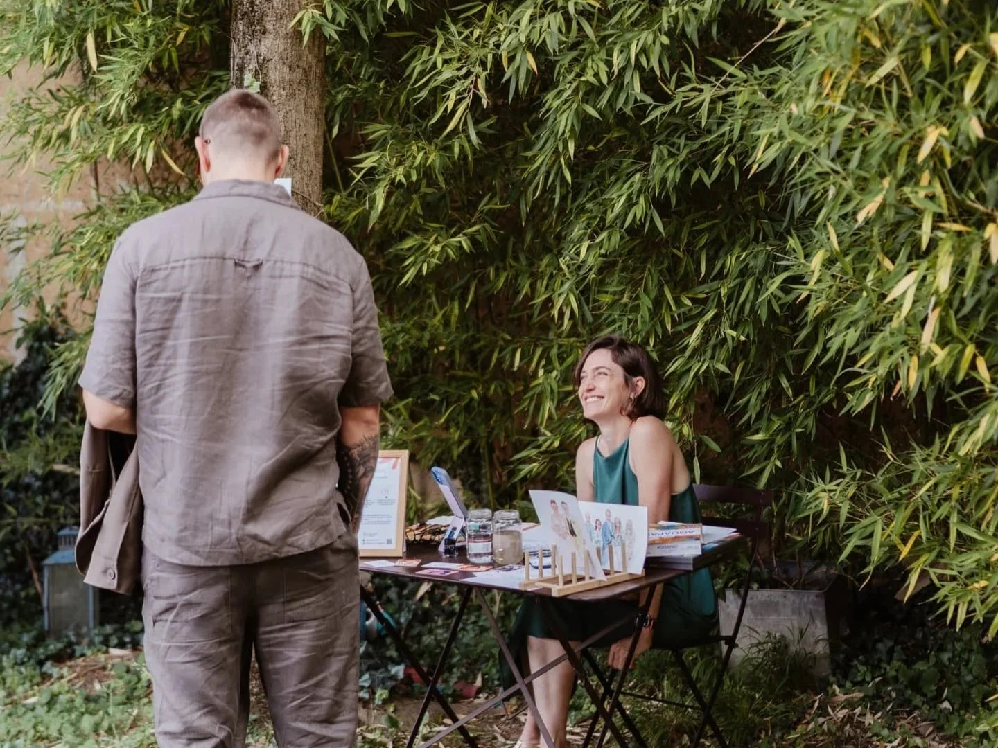 Une femme souriante assise à une table en extérieur devant un stand avec des affiches et des livres, rencontrant un homme dont le dos est visible, dans un environnement gazonné avec des arbustes et un arbre.