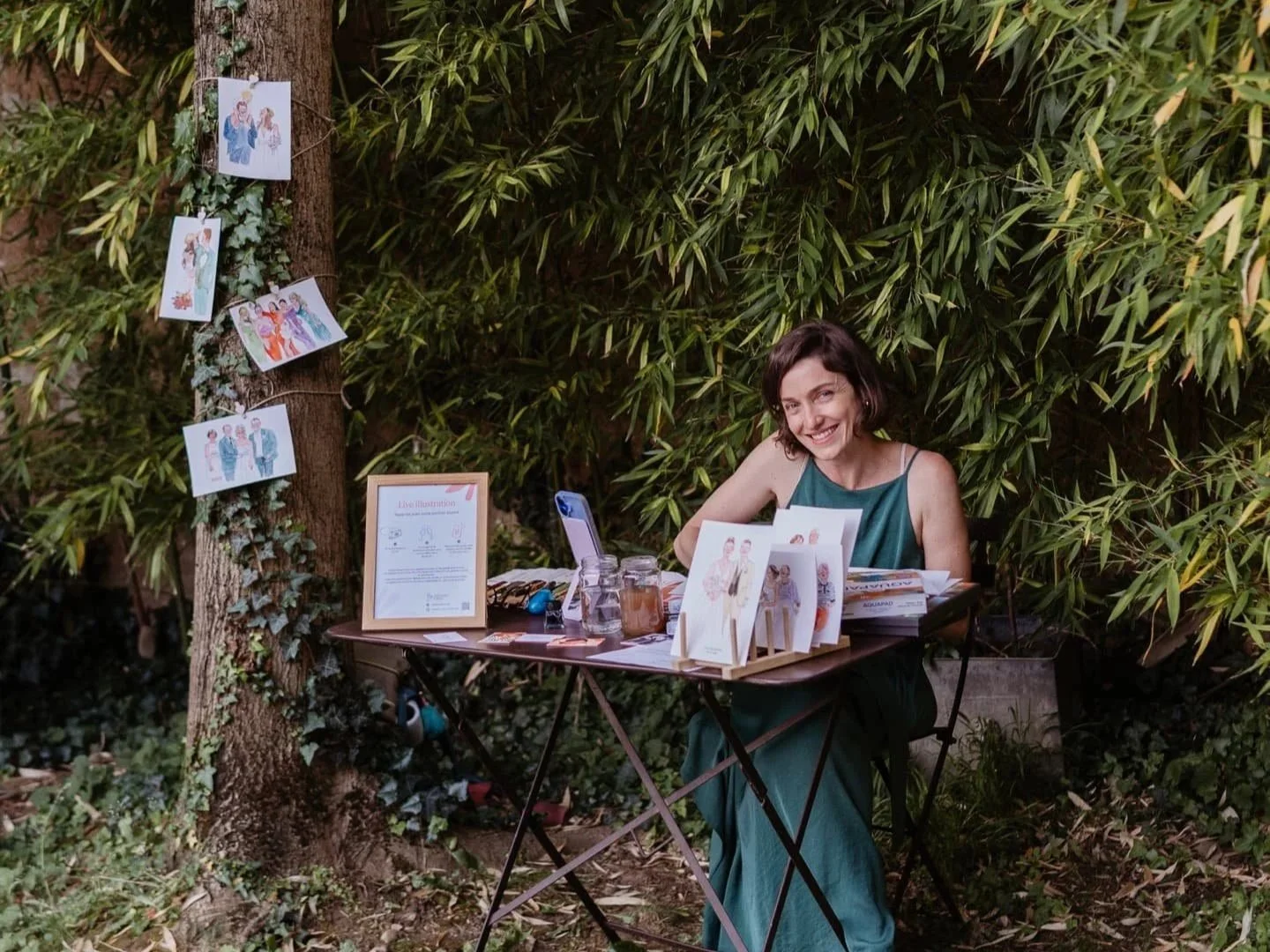 Une femme souriante assise à un stand d'illustrations dans un espace extérieur entouré de feuillage dense.