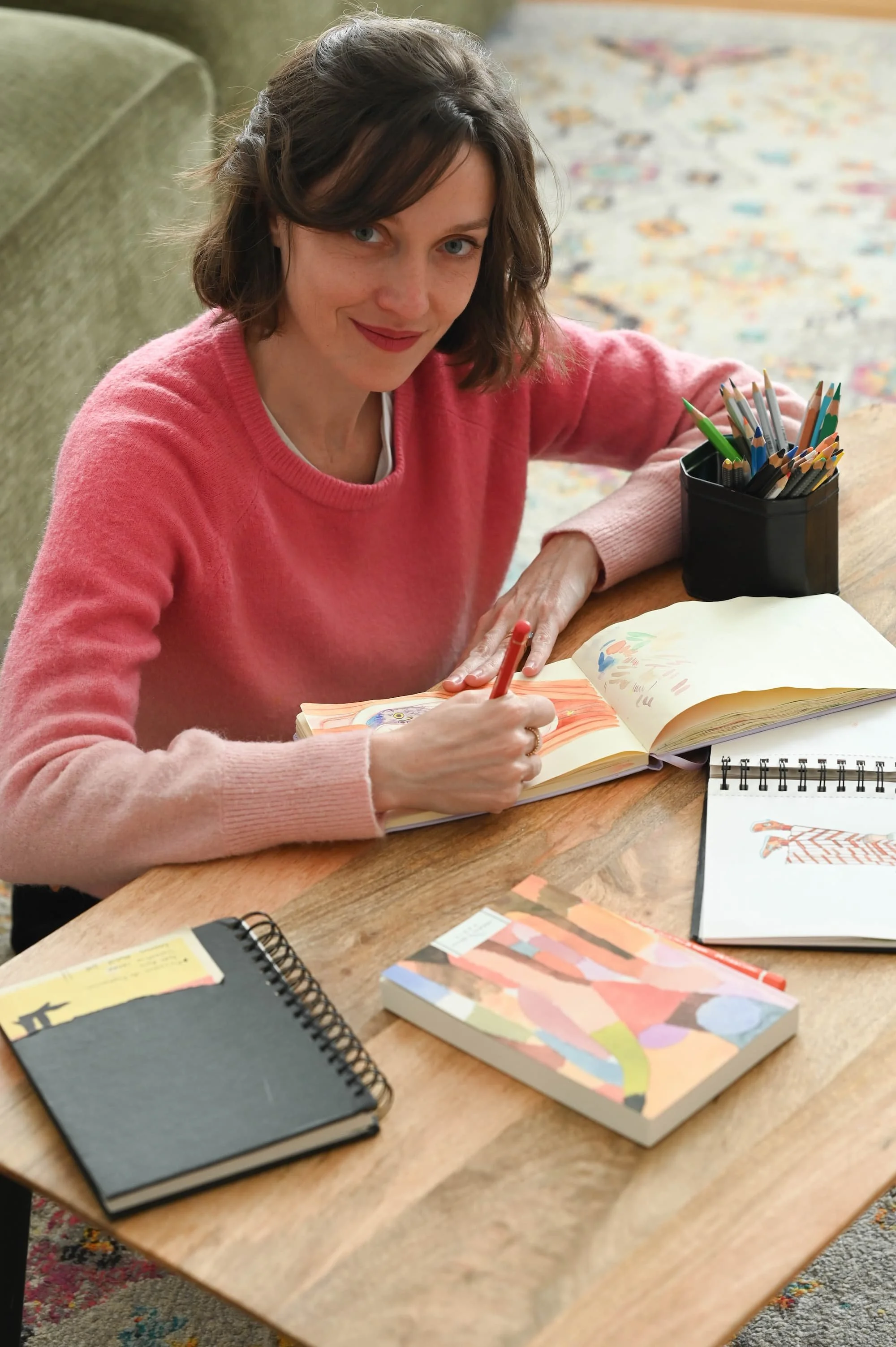 Une femme souriante, assise à une table en bois, en train de dessiner dans un carnet avec des crayons de couleur, entourée de plusieurs carnets, un pot à crayons et un tapis coloré en arrière-plan.