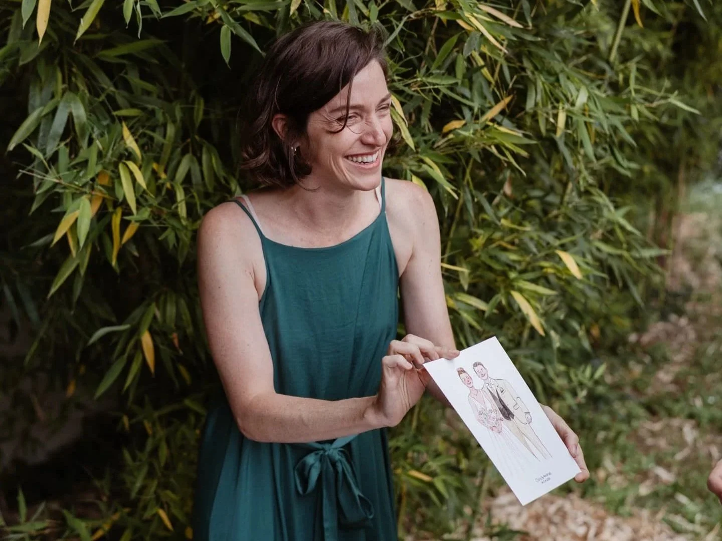 Femme souriante en robe verte tenant une illustration de couple lors d'un événement en plein air.
