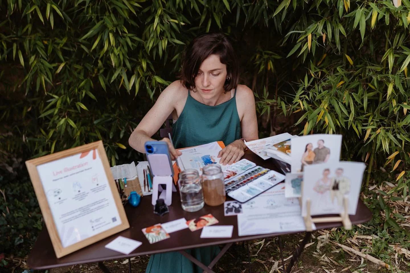Une femme assise devant une table en plein air, entourée de feuilles vertes, en train de faire de la peinture ou du dessin, avec des fournitures artistiques et des photos sur la table.
