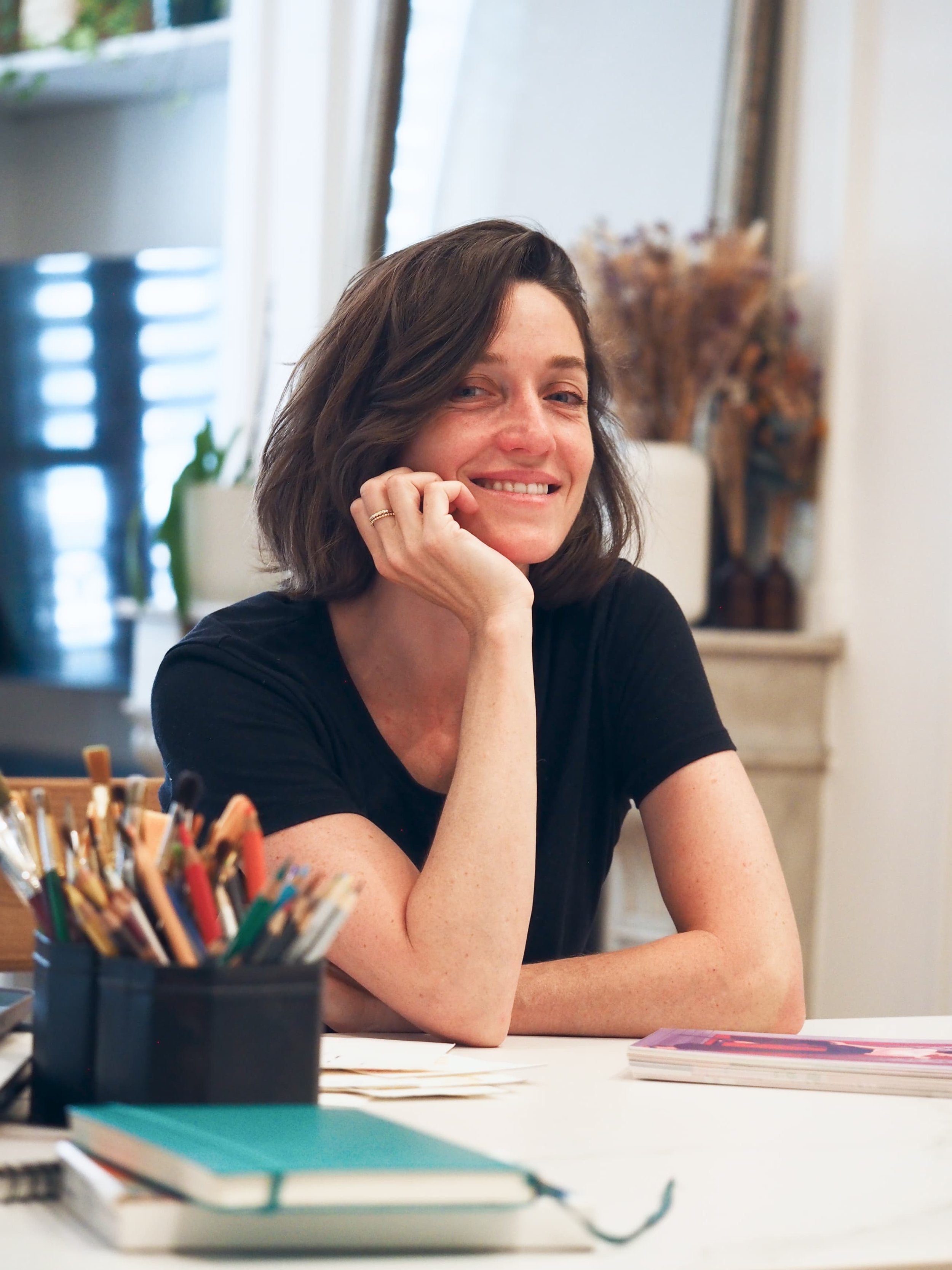 Valentine Carre illustratrice et directrice artistique souriante assise à une table avec des livres et des crayons de couleur en premier plan.