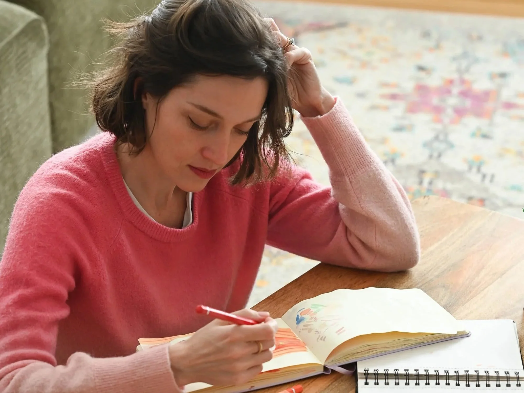 Femme assise à une table, portant un Pull rose, écrivant dans un carnet avec des crayons de couleur ouverts devant elle.