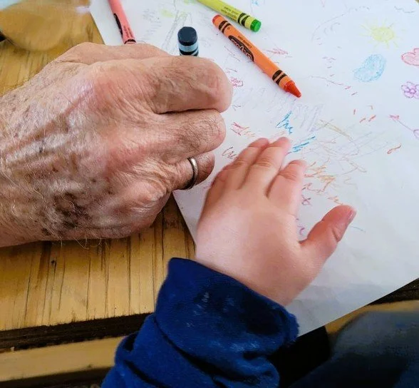 Close-up of a child's hand drawing with crayons on a piece of paper while an elderly person's hand holds the paper steady on a wooden table.