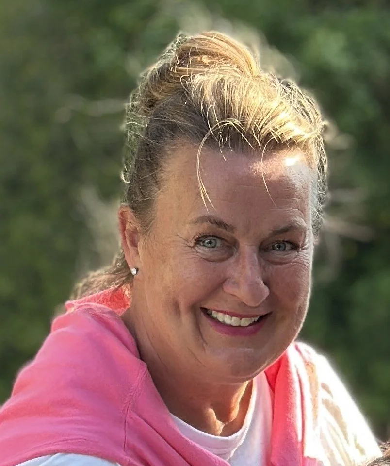 A smiling woman with blonde hair in a bun, wearing a pink garment outdoors with green trees in the background.