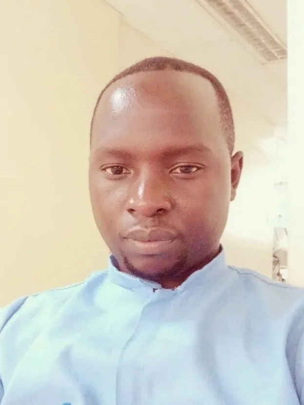 A young man with short hair and a slight beard wears a light blue shirt and looks directly at the camera. The background is a plain wall and part of an indoor setting.