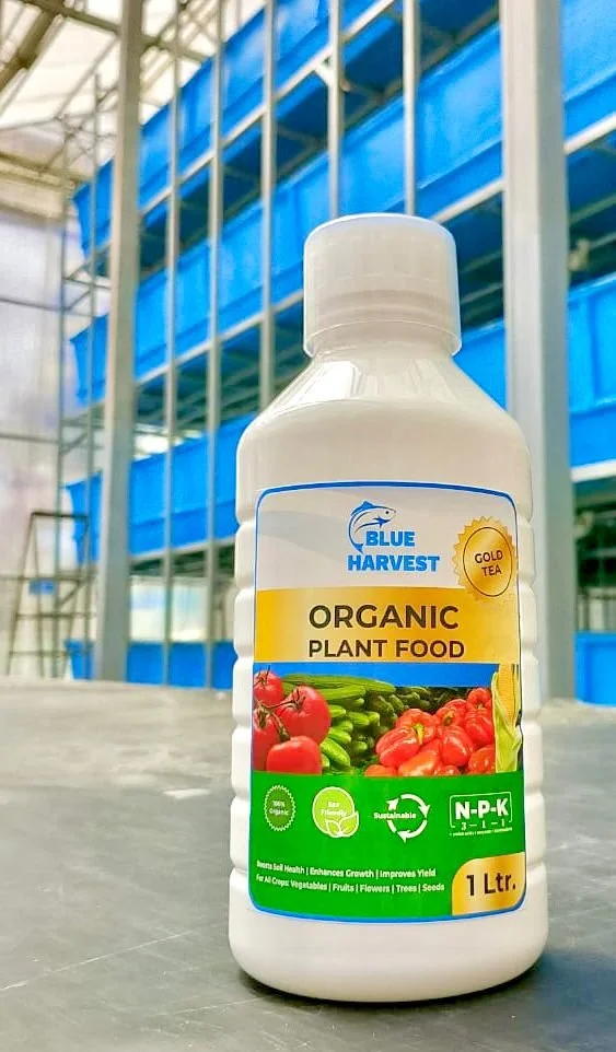 A white bottle labeled Blue Harvest Organic Plant Food, placed on a table in a greenhouse with blue plastic containers and metal shelves in the background.