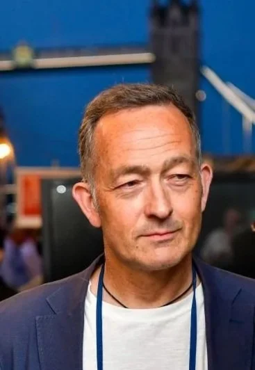 A middle-aged man with short dark hair and a slight smile stands indoors at an event, wearing a dark blazer over a white shirt.