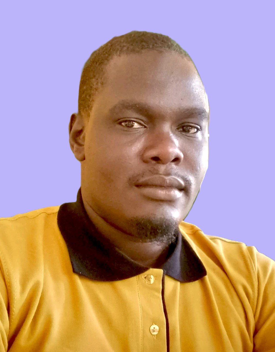 A young man with short hair and a goatee wearing a yellow shirt with a black collar, looking into the camera against a purple background.