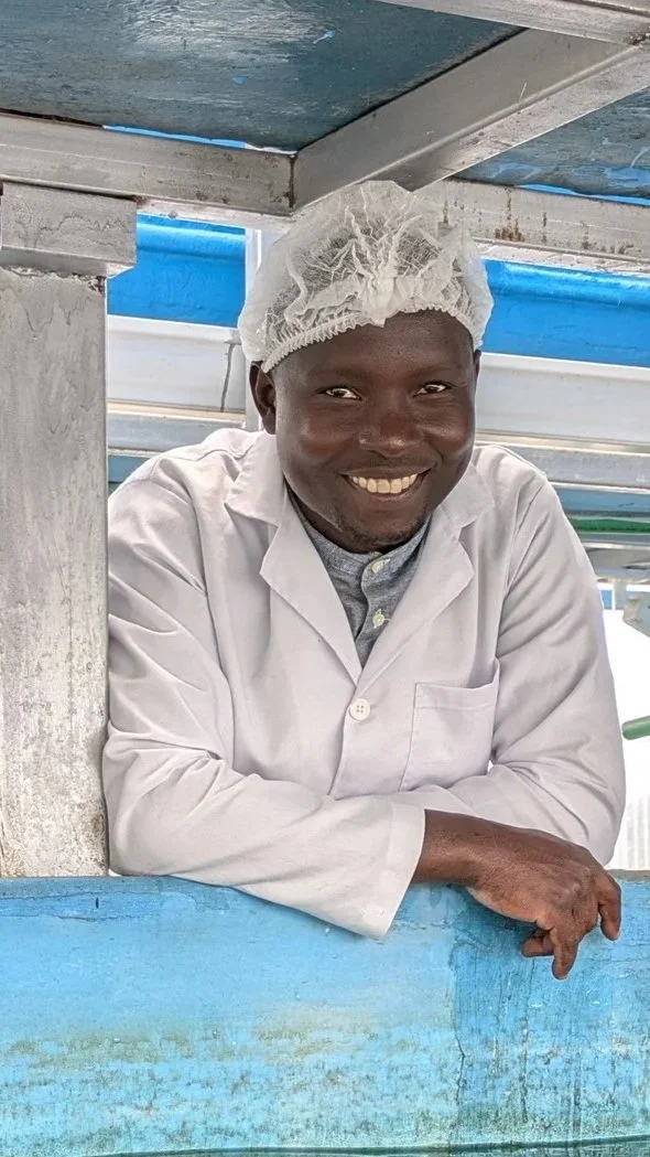 Smiling man in a white lab coat and a hairnet leaning on blue-painted wooden railing in a clean indoor setting.