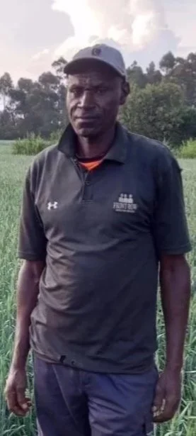 A woman and a man standing in a green field with tall grass, trees in the background, and a partly cloudy sky. The woman wears sunglasses, a blue shirt, and a long black skirt. The man wears a cap, a black polo shirt, dark pants, and rubber boots.