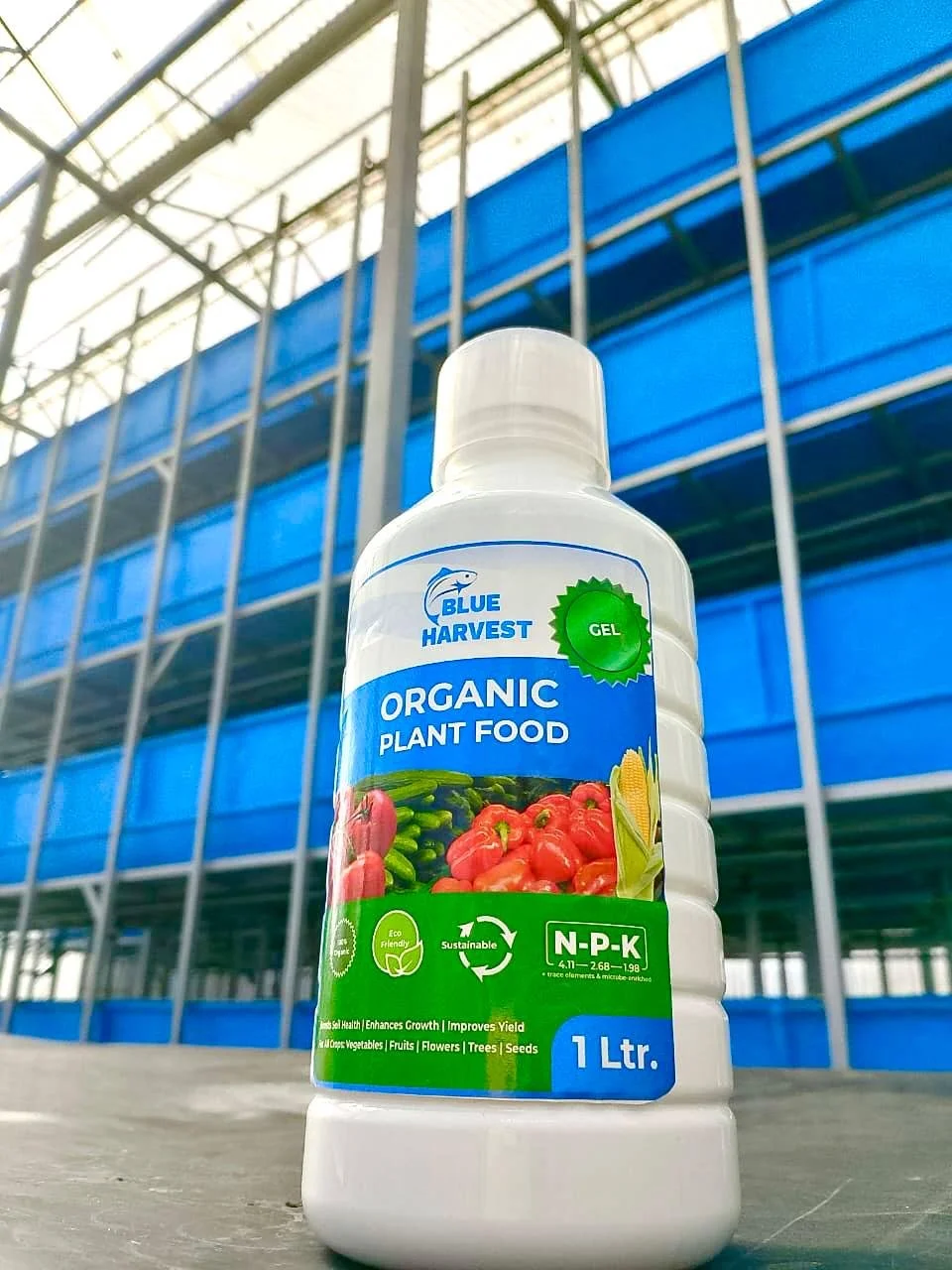 A 1-liter bottle of Blue Harvest organic plant food gel with images of vegetables and fruits on the label, placed in front of a multi-story building with blue and green accents.