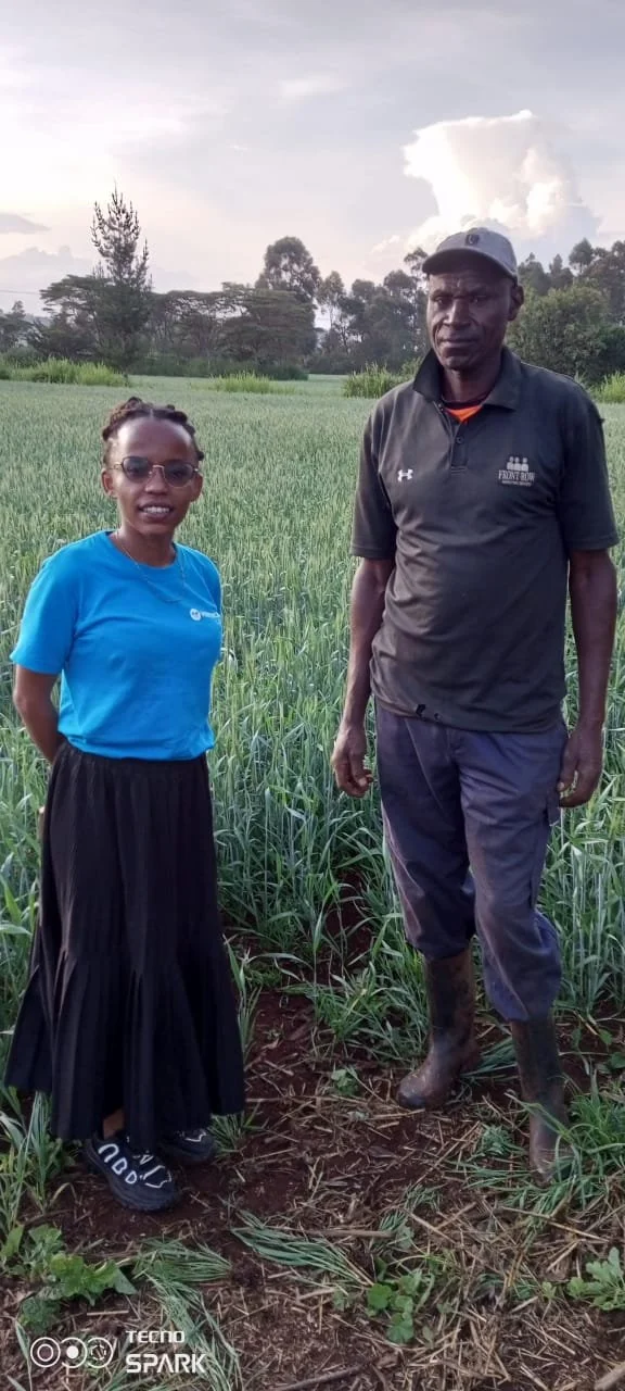 A woman and a man standing in a green field with tall grass, trees in the background, and a partly cloudy sky. The woman wears sunglasses, a blue shirt, and a long black skirt. The man wears a cap, a black polo shirt, dark pants, and rubber boots.
