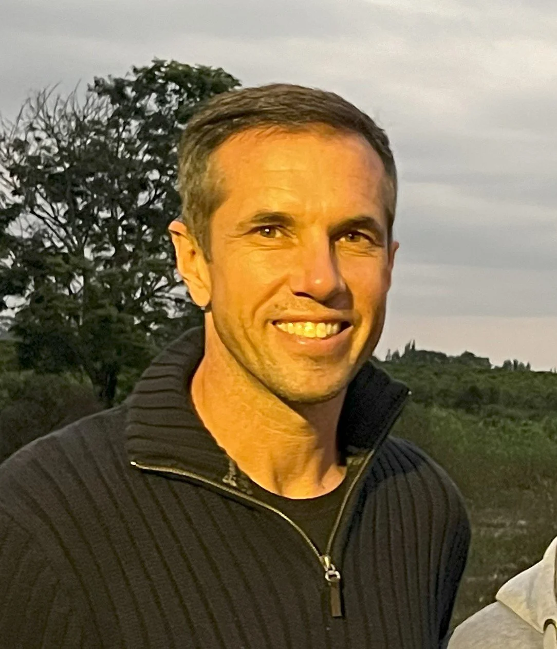 A man smiling outdoors with a tree and cloudy sky in the background.
