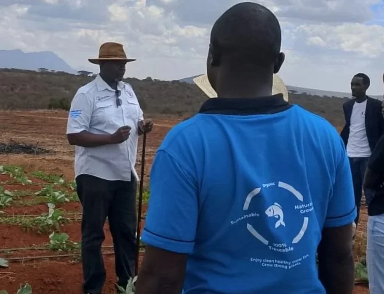 A group of people standing in an agricultural field with a woman holding a walking stick, wearing a wide-brimmed hat, and others wearing t-shirts and jackets, under a partly cloudy sky.
