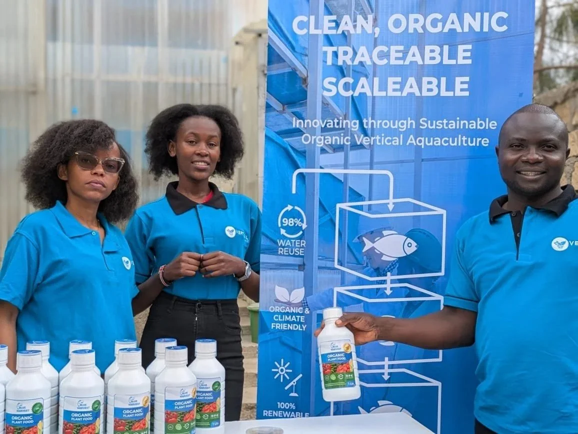 Three people standing behind a table with bottles of organic plant food, beside a large blue banner with text about sustainable organic vertical aquaculture.