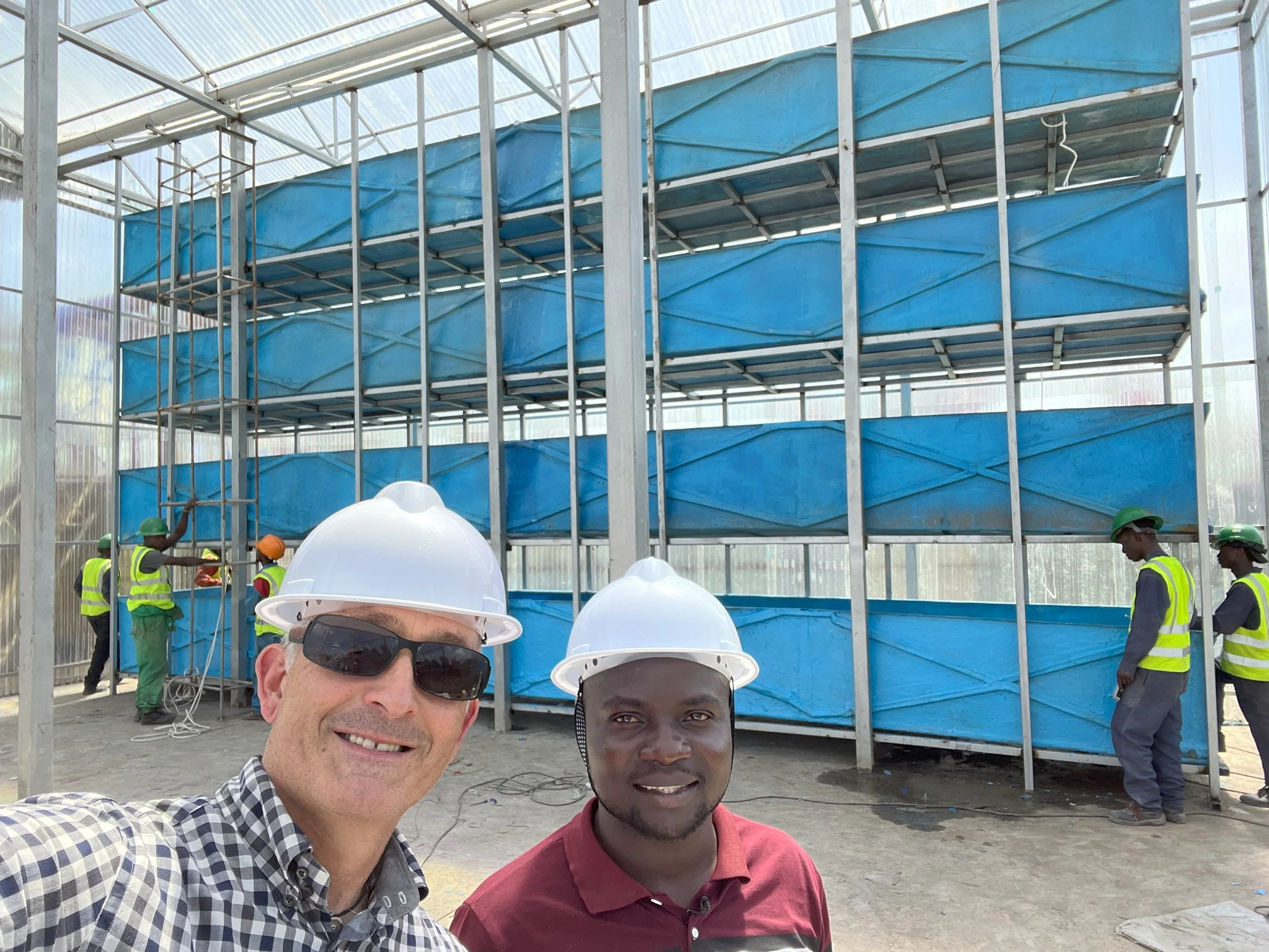 Two men wearing white hard hats taking a selfie in front of a construction site with several workers installing metal and blue panels on a large structure.