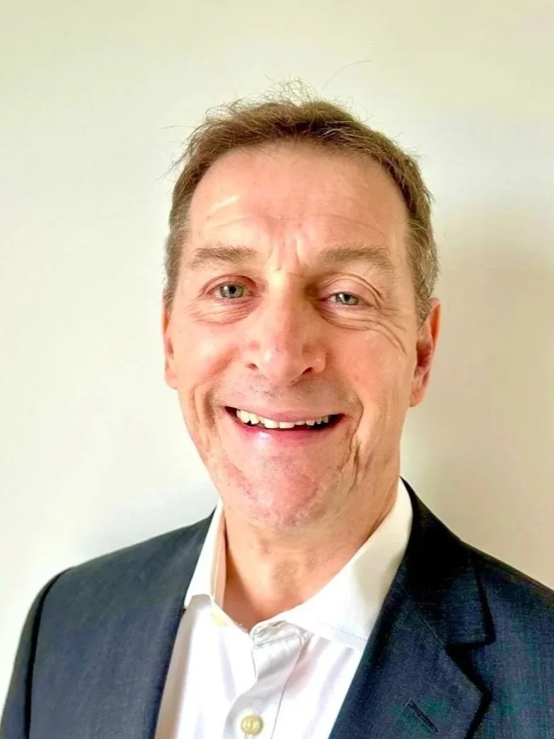 A middle-aged man with light skin, short light brown hair, and blue eyes, smiling and wearing a white shirt and a dark suit jacket against a plain light-colored background.