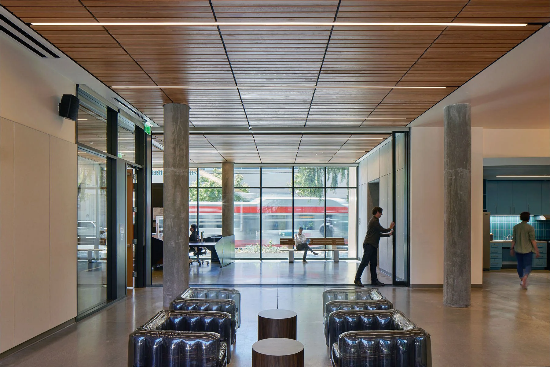 Modern waiting area inside a building with large glass windows, cushioned chairs, and people sitting and standing.