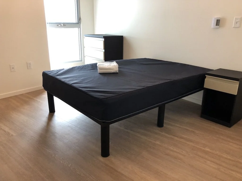 A bedroom with a black bed frame, black mattress, white folded towel on the bed, light-colored wooden floor, a black and white dresser near the window, and a small black nightstand beside the bed.