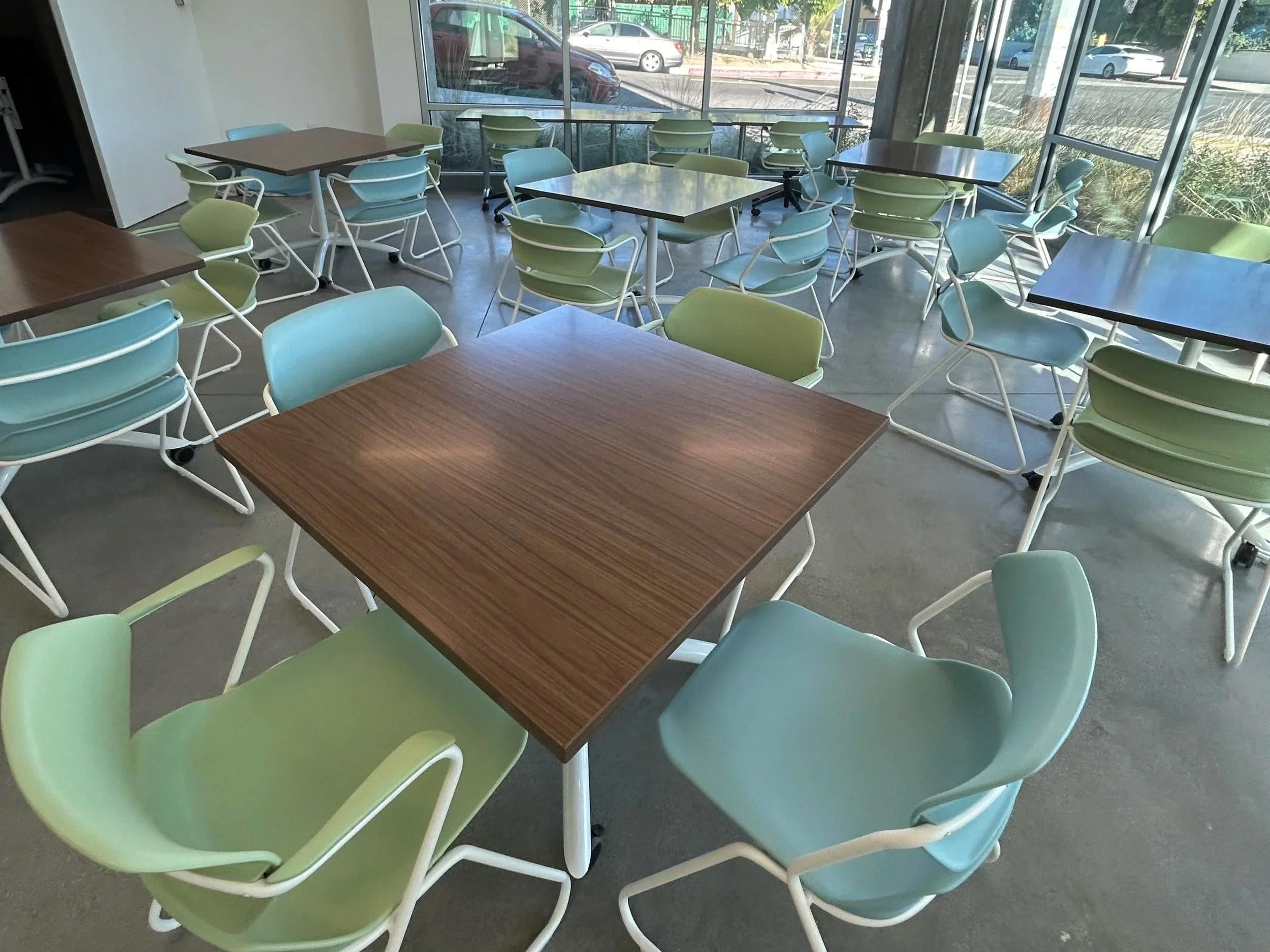 Empty indoor restaurant or cafe with light blue and green chairs and dark wooden tables, large windows showing a parking lot outside, and polished concrete floor.